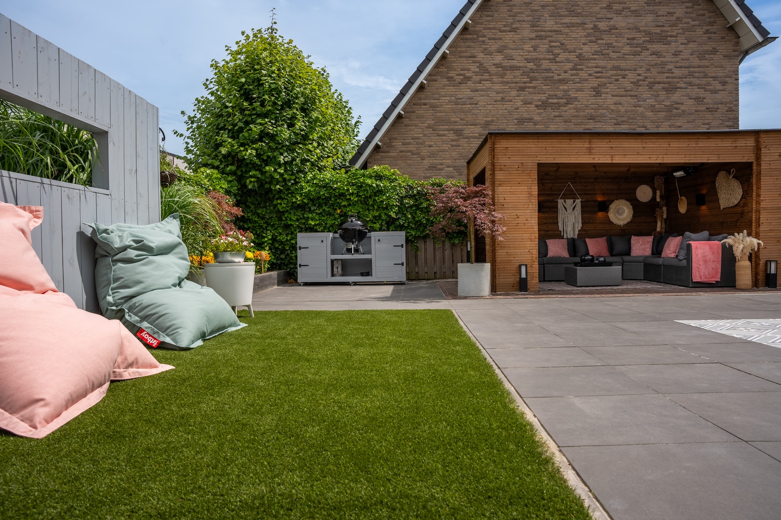 De achtertuin heeft een grillstation met een strak, modern design tegen een achtergrond van weelderige groene planten en bloemen. De ruimte is goed georganiseerd, met een mix van kunstgras en betegelde gebieden.