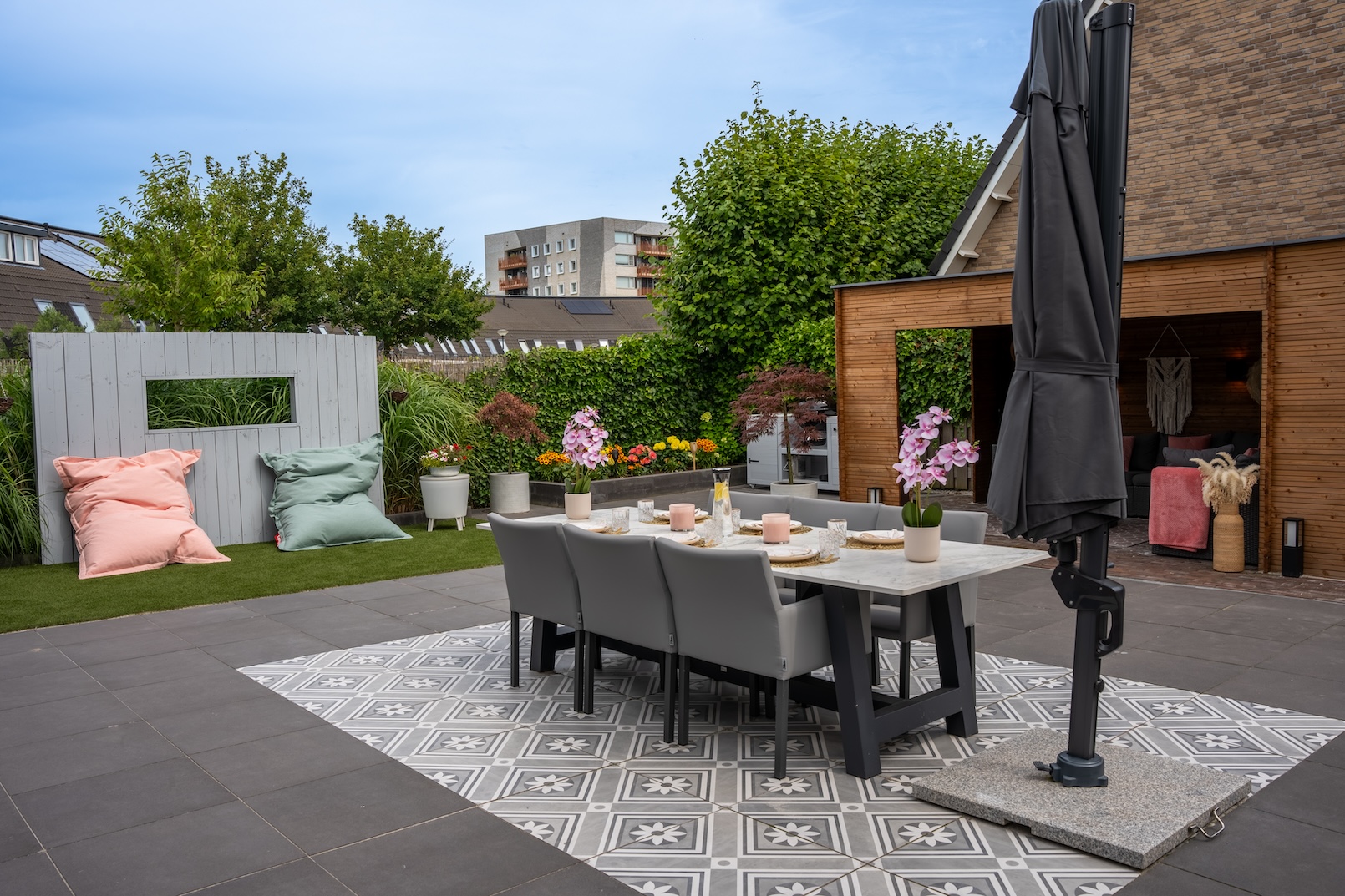 Tuinrenovatie - Een close-up van de gezellige zithoek in de houten pergola. De ruimte is ingericht met zwarte en roze kussens, hangende geweven decoraties en planten in potten.