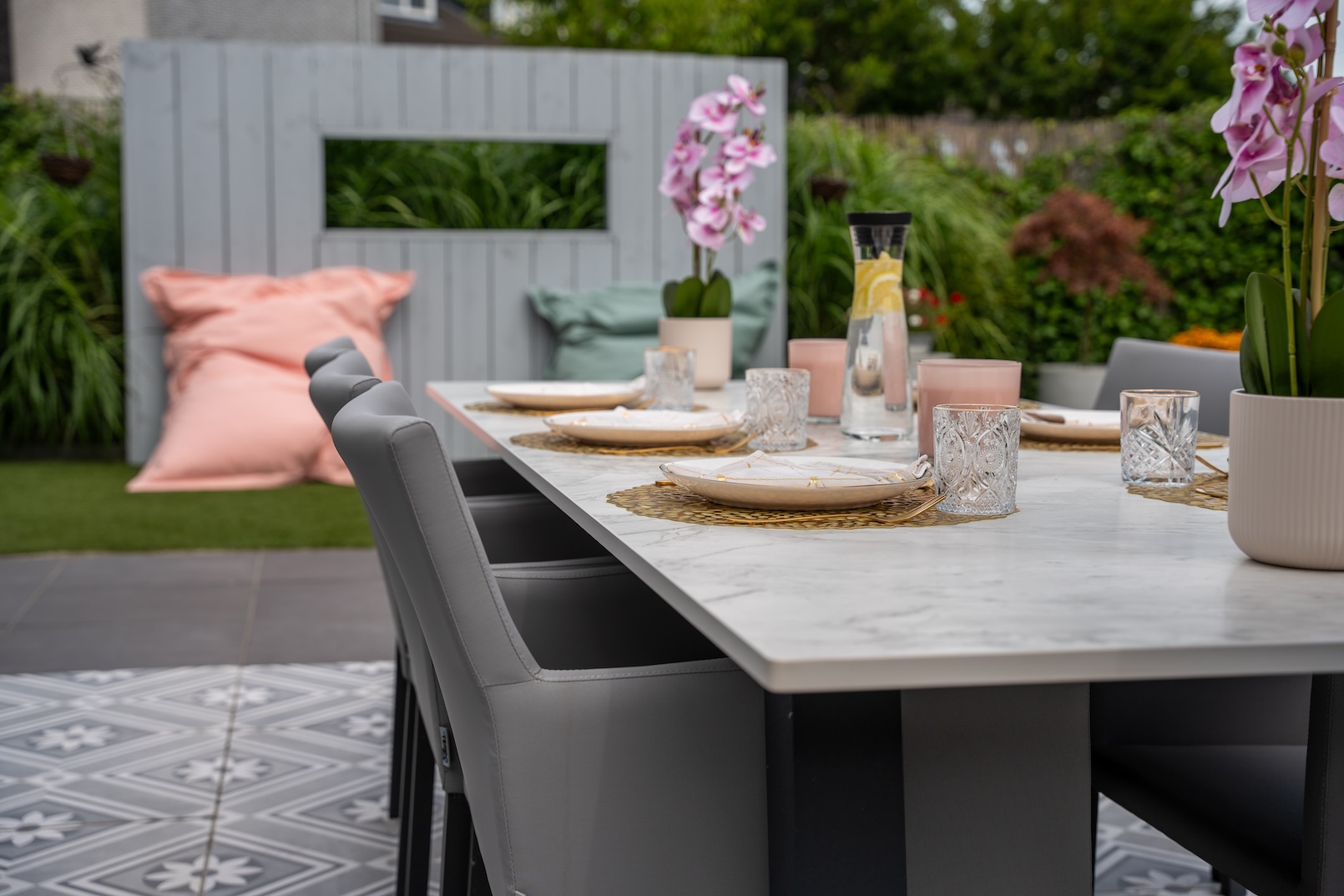 Een buiteneetgedeelte met een strak grijs tafel en stoelen set op een betegeld terras. Het eetgedeelte is versierd met roze bloemen en de achtertuin is omgeven door een zwart houten hek met verschillende planten.