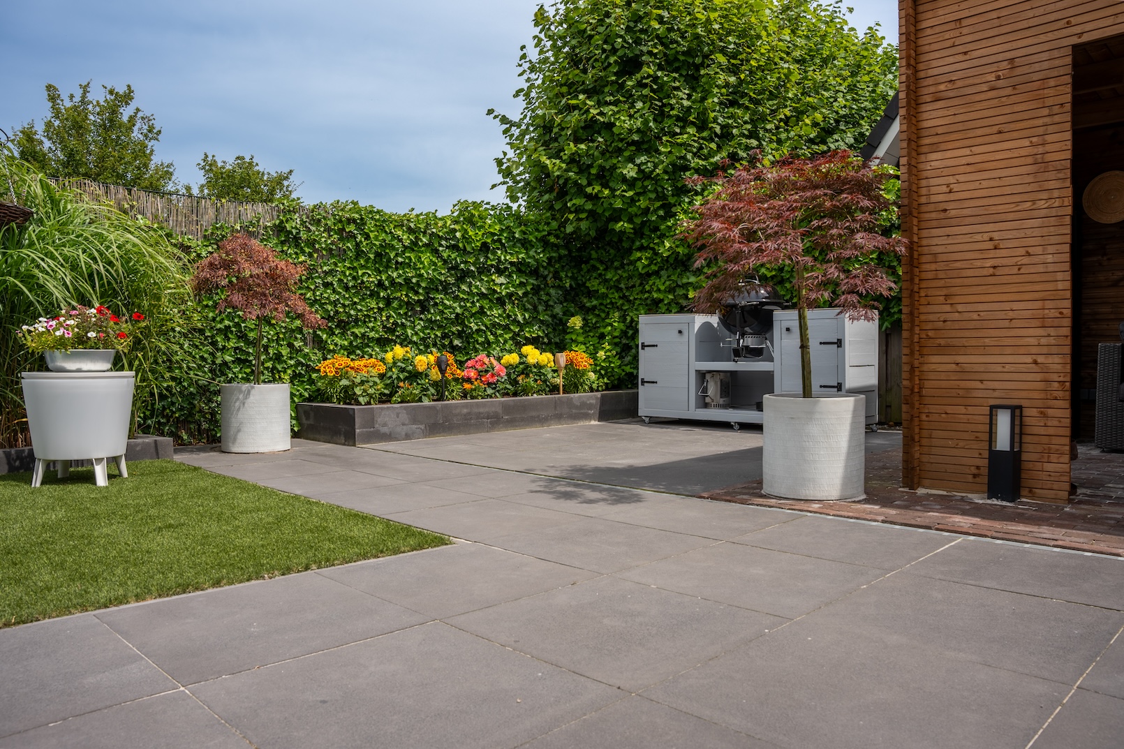 Een ander perspectief van de achtertuin, waarbij de continuïteit van het betegelde gebied naar de eetruimte en de zithoek in de houten pergola te zien is. Het kunstgras voegt een levendige touch toe aan het algehele ontwerp.