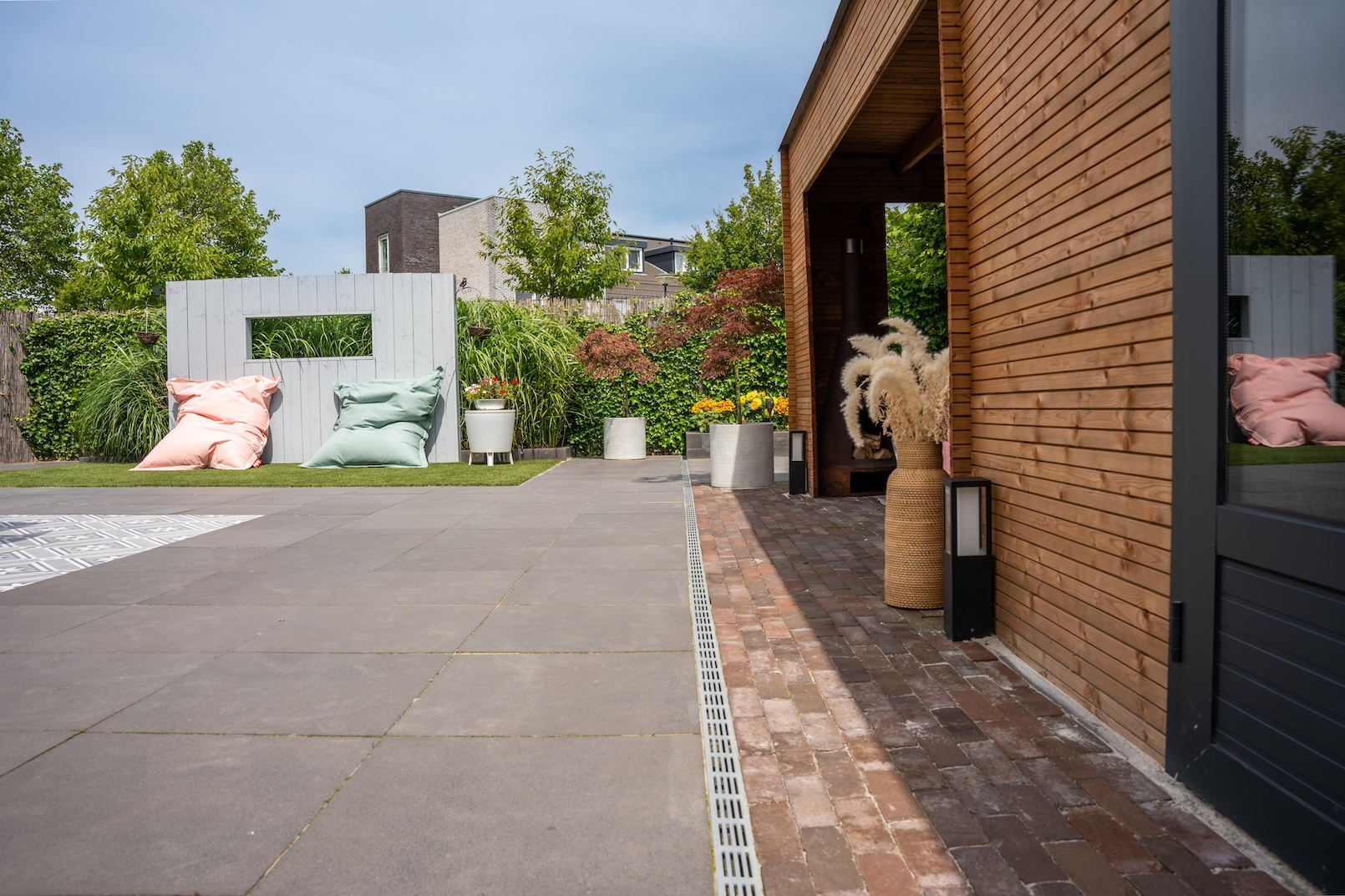 Een ander perspectief van de achtertuin, waarbij de continuïteit van het betegelde gebied naar de eetruimte en de zithoek in de houten pergola te zien is. Het kunstgras voegt een levendige touch toe aan het algehele ontwerp.